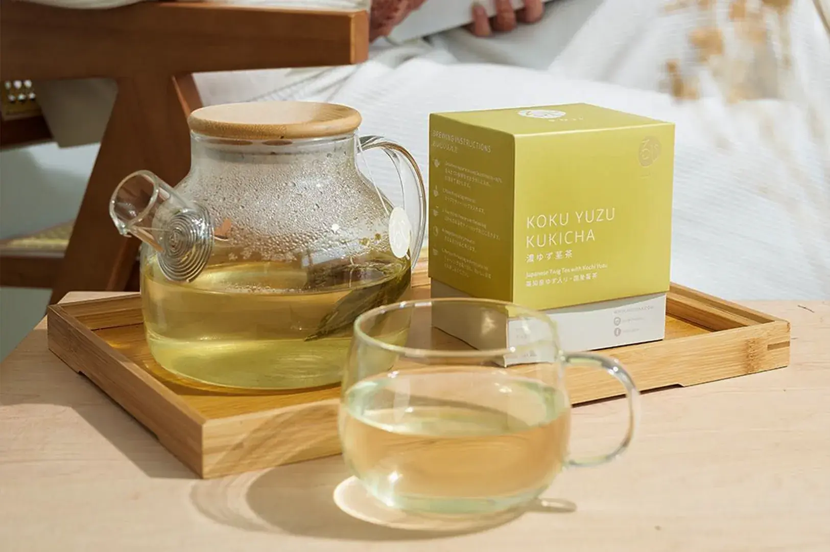 A serene, brightly lit tea-brewing setup arranged neatly on a shallow wooden tray over a light-colored tabletop. On the left sits a clear, round glass teapot with a light wood lid, half-filled with freshly steeped, pale yellow-green tea containing a single submerged tea sachet. To the right of the teapot stands a square, light-chartreuse product box labeled "Koku Yuzu Kukicha Japanese Twig Tea with Kochi Yuzu," with brewing instructions printed in white on its side panel. In the foreground rests a matching clear glass teacup filled with the golden-hued brew, casting a soft shadow on the surface.