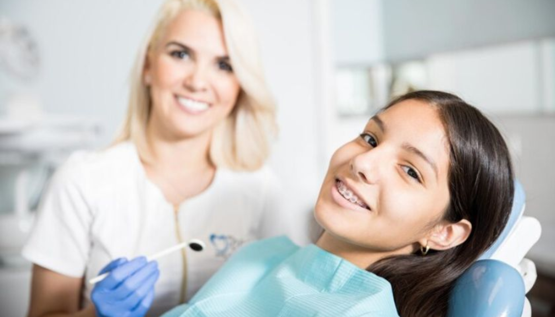 Smilende jente med tannregulering sitter i tannlegestol, mens kvinnelig tannlege med hansker holder et tanninstrument ved siden av.