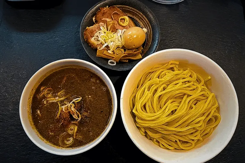 A bowl of thick noodles sits next to a bowl of rich, brown dipping sauce topped with green onions. In the background, there is a dish with sliced pork, boiled egg, and vegetables. A water glass is placed nearby on the black wooden table, creating a warm, inviting dining scene.