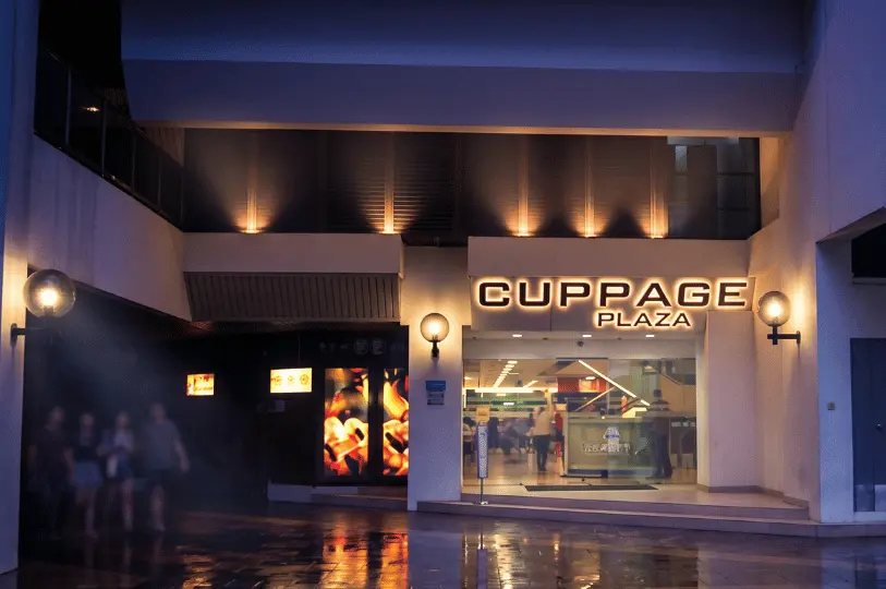 Exterior night view of the Cuppage Plaza building in Singapore with illuminated signage and blurred pedestrians walking by.