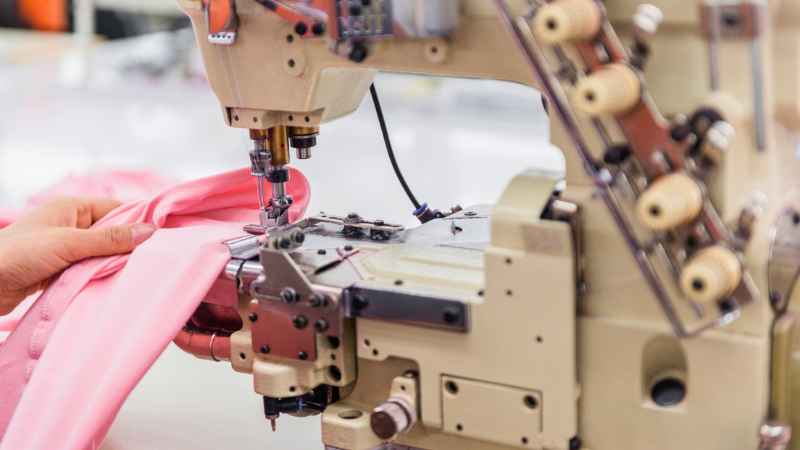 A sewing machine stitches pink fabric held by hands.