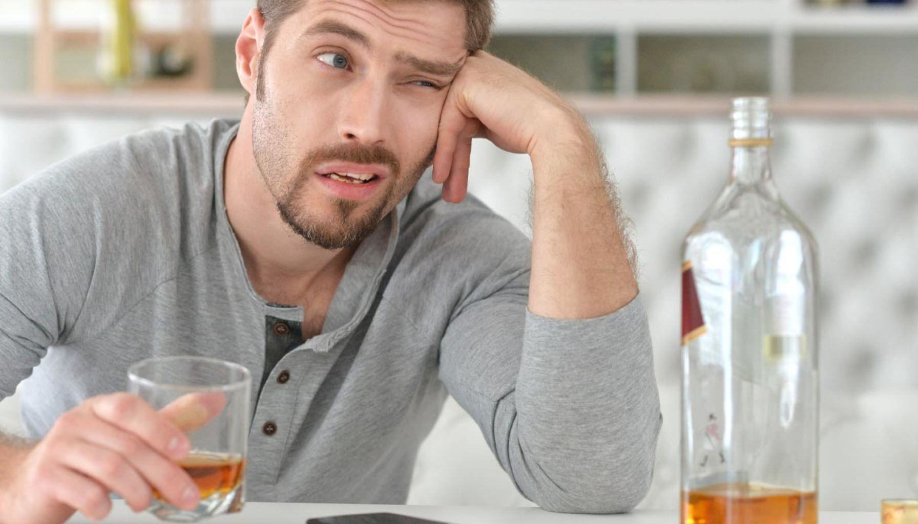 Alkohol etter tanntrekking Hvorfor du bør vente 12 En ung mann sitter sliten med et glass alkohol, halvfull flaske foran seg og et urolig uttrykk i ansiktet.