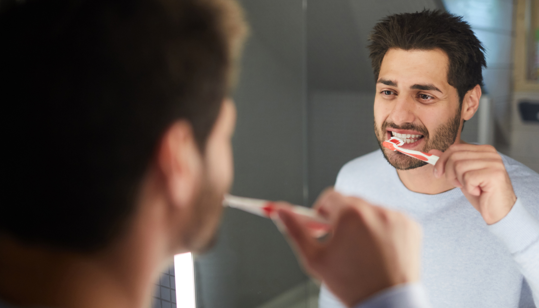 Mann pusser tennene foran speilet på badet med rød tannbørste og ser konsentrert ut.