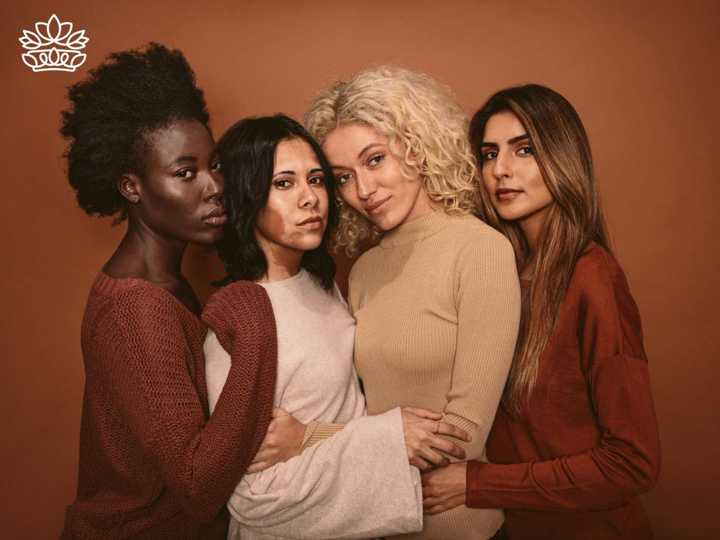 Four women of different ethnicities closely embracing, portraying strong and diverse beauty in warm-toned sweaters. Perfect for celebrating connections with gift hampers including body lotion, milk chocolate, and white chocolate from the Gifts Boxes for Women Collection. Fabulous Flowers and Gifts