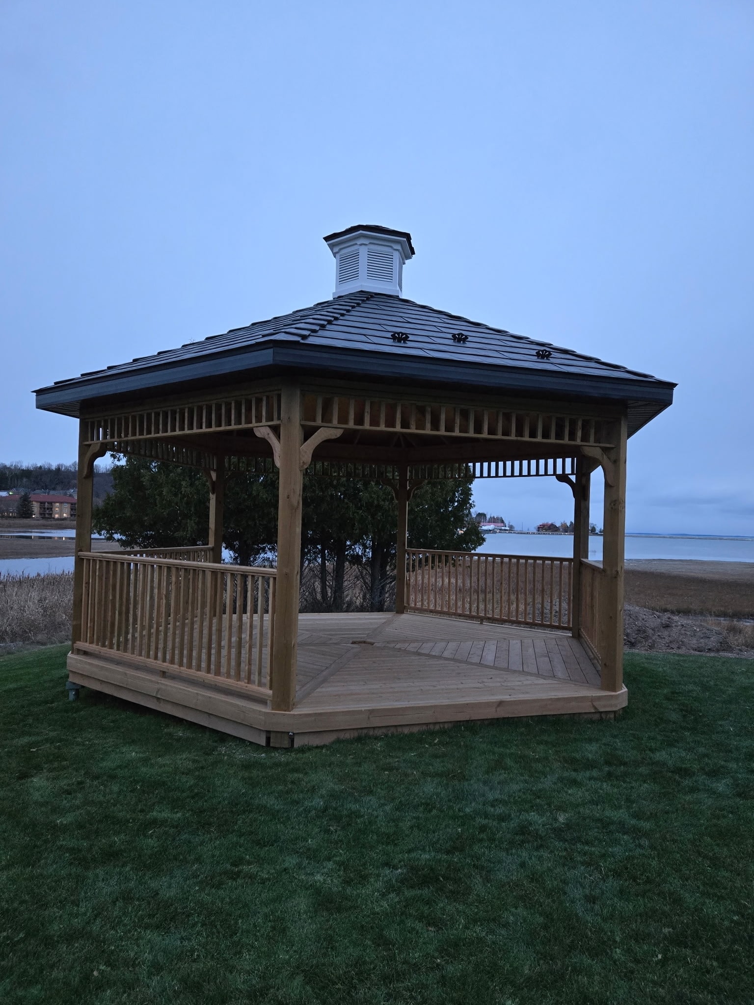 A wooden gazebo with a dark metal roof overlooks the water, standing on a sprawling grassy area.
