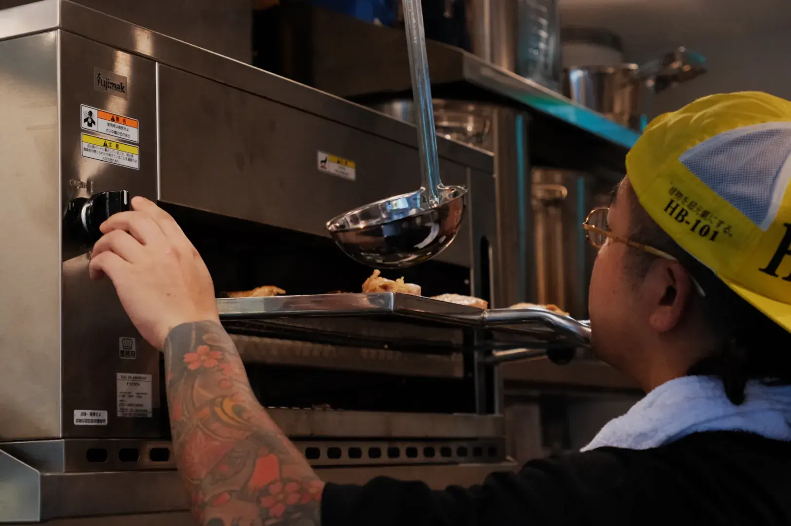 A person with tattoos on their arm adjusting a temperature dial on a commercial oven while holding a ladle.