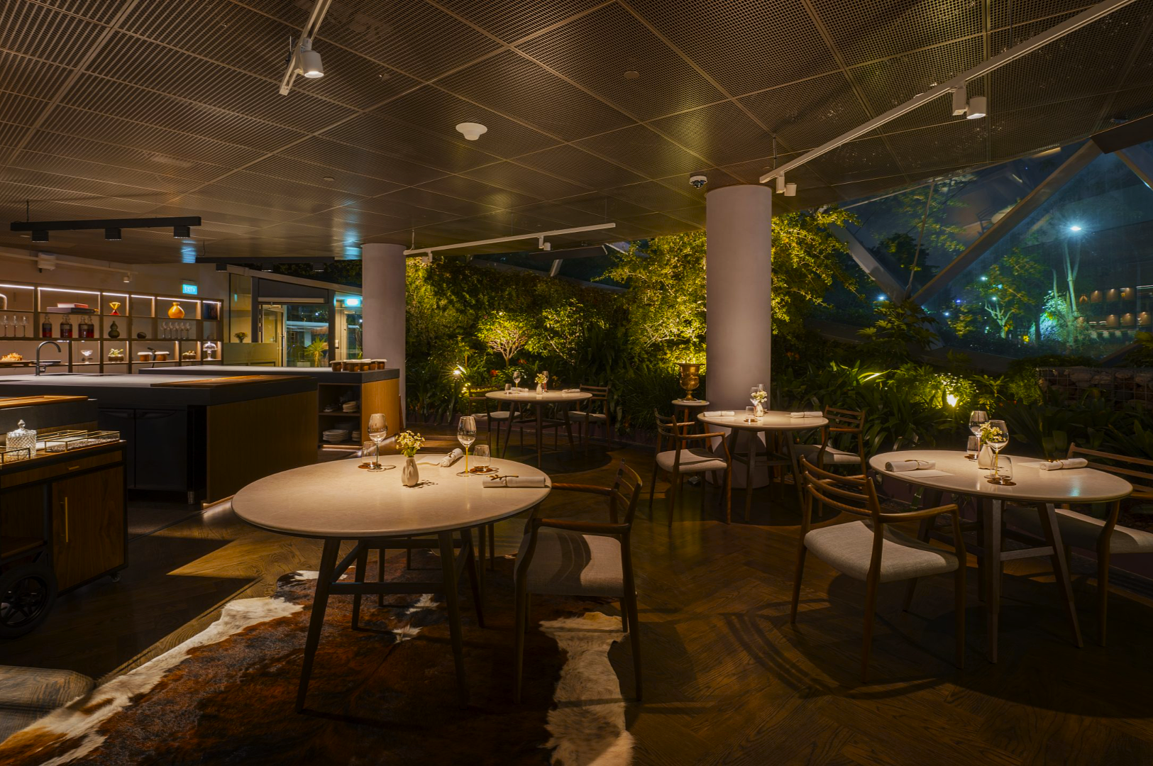 Elegant restaurant with round tables and wooden chairs set for dining. Ambient lighting, indoor greenery, and a calm, cozy atmosphere prevail.