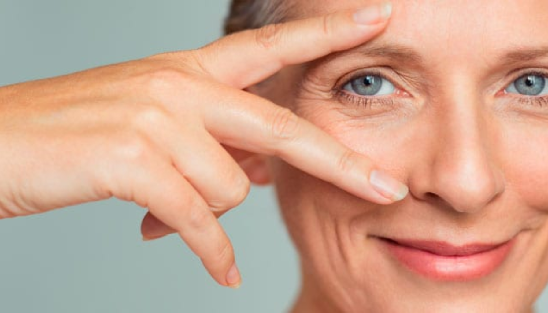 Moden kvinne smiler rolig mens hun holder fingrene i V-form rundt det ene øyet, med fokus på sunn hud og blå øyne.
