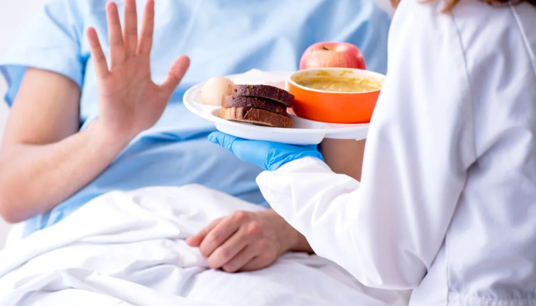 Pasient på sykehus seng avviser matbrett med suppe, brød og eple servert av helsepersonell.