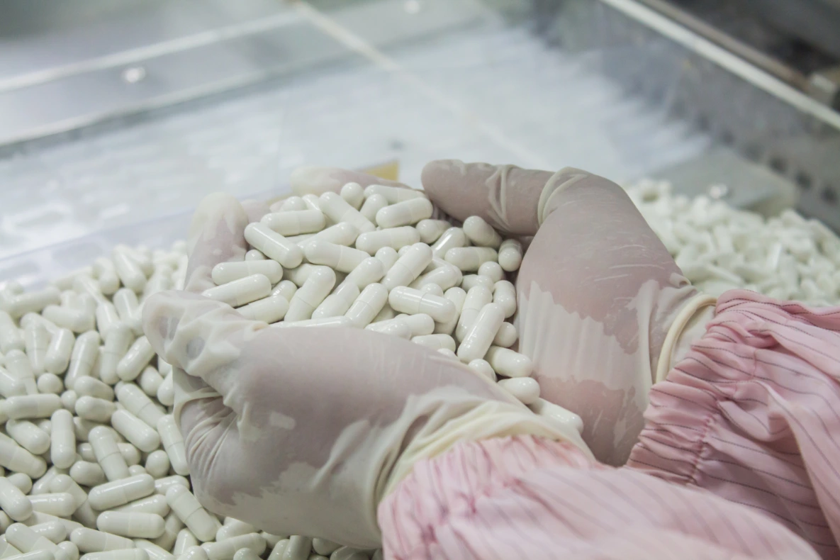 Hands in gloves holding white capsules with a large quantity of the capsules behind them.