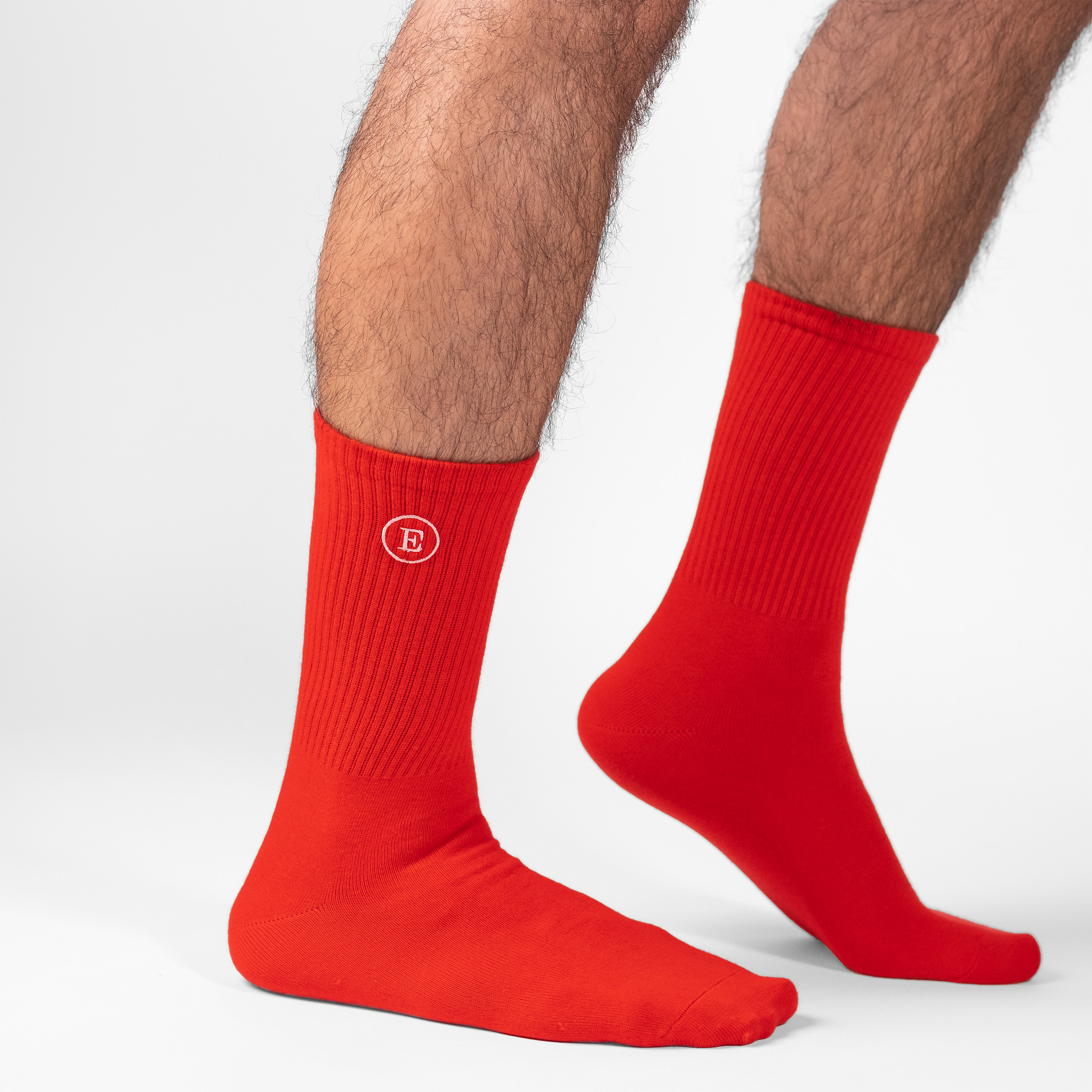 A male model wearing a pair of personalised red crew socks with the initial 'E'embroidered in white thread on the outside leg of the sock.