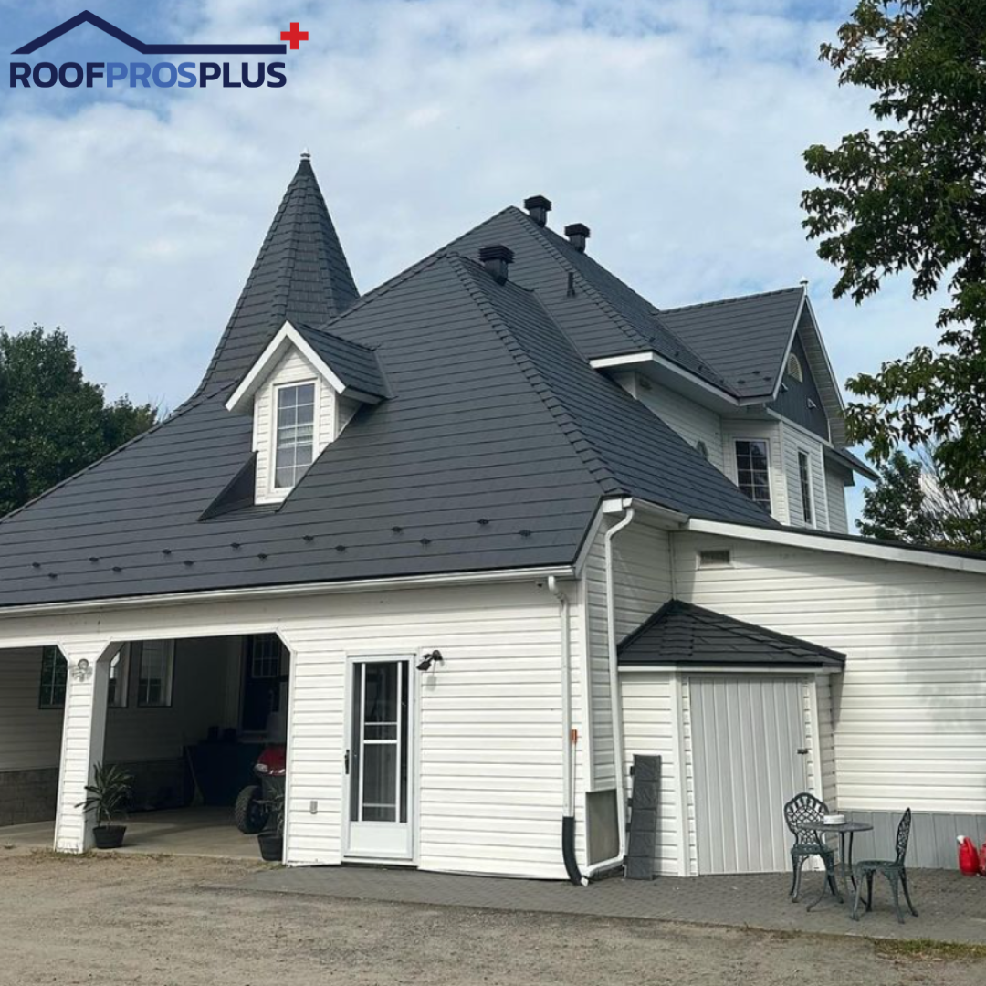 A white house with a steep, dark grey metal roof, featuring multiple peaks and a dormer. The wide garage is open, with a small patio table outside.