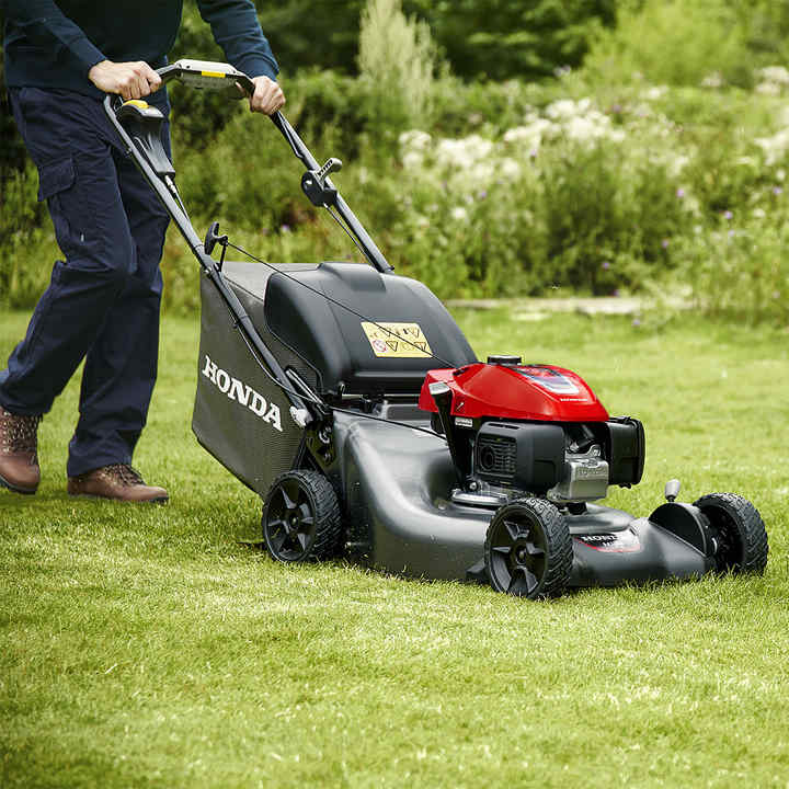 A person is operating a Honda lawn mower with a red engine and black body on a well-maintained green lawn. The mower has a grass catcher at the back, and the user is wearing dark pants and brown shoes. The background features lush greenery and flowering plants.