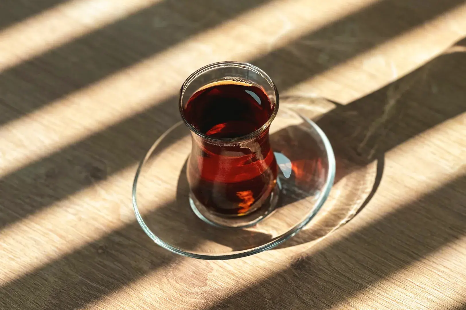 Tulip-shaped glass of dark tea on a clear saucer resting on a light wooden table illuminated by diagonal window blind shadows.