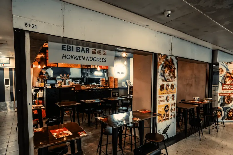 Storefront of Ebi Bar Hokkien Noodles located at unit B1-21 inside a dimly lit basement corridor of Cuppage Plaza, with empty tables and chairs set up for diners.