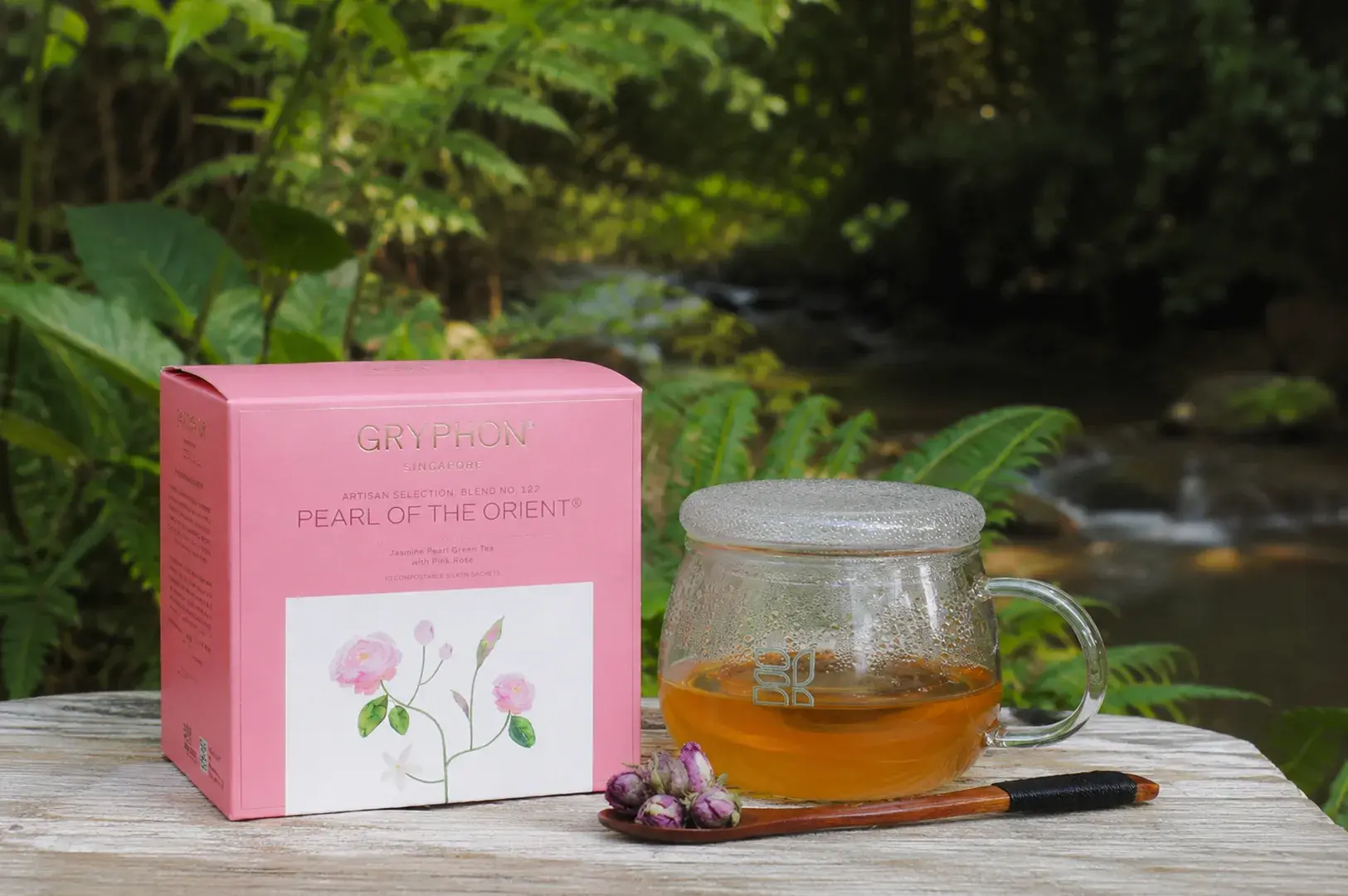 A pastel pink box of Gryphon "Pearl of the Orient" tea is positioned on a rustic, weathered grey wooden table, set against a beautiful background of a flowing forest stream and dense green wilderness. A round, clear glass teacup with a matching glass lid sits next to the box, filled with a translucent golden-orange tea. A dark wooden spoon with a black-cord-wrapped handle lies in front of the teacup, cradling a small cluster of dried pink rosebuds that complement the floral theme illustrated on the tea packaging.