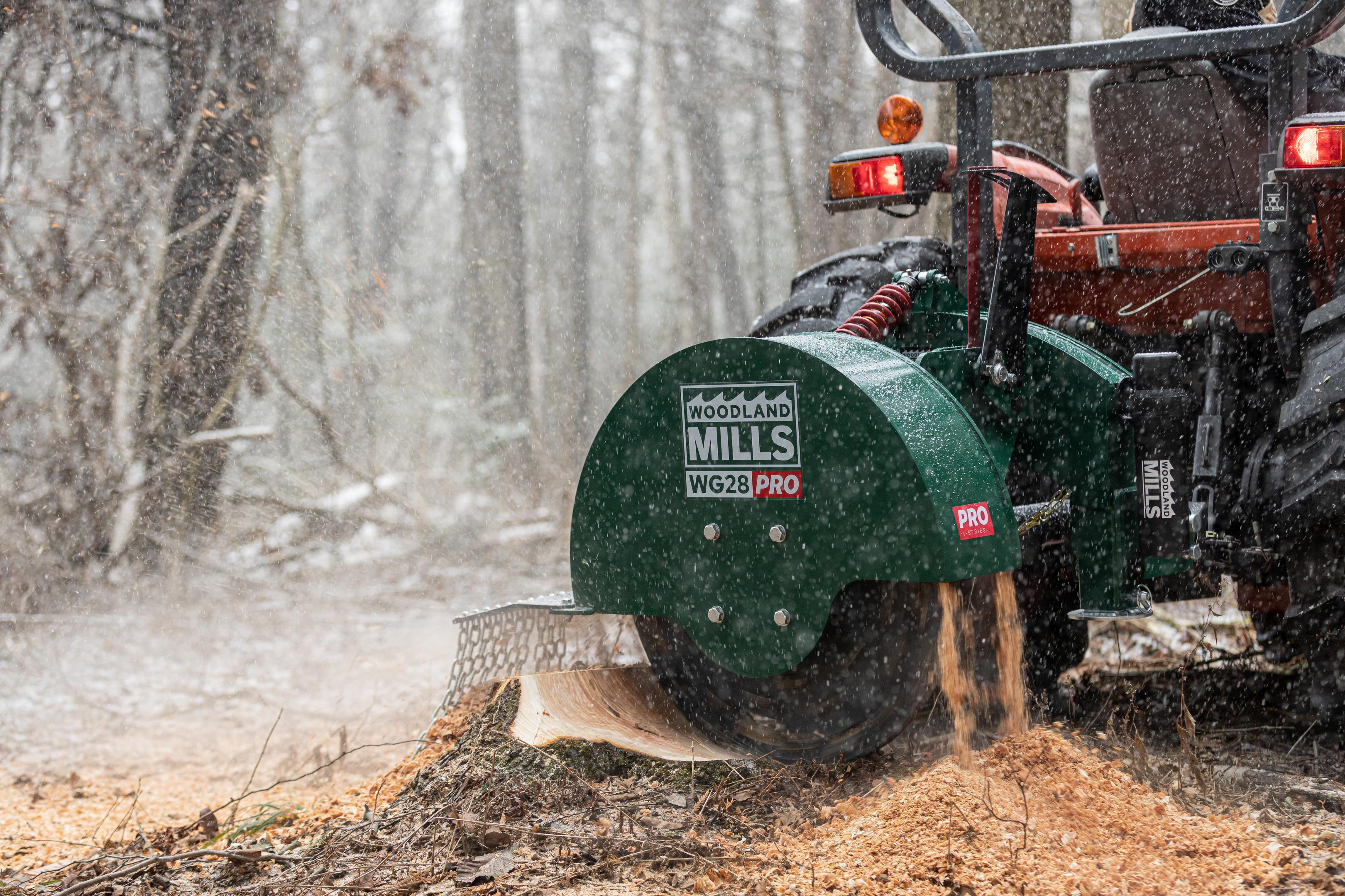 A Woodland Mils WG28 PRO Stump Grinder attached to a tractor in a snowy forest grinding a stump.
