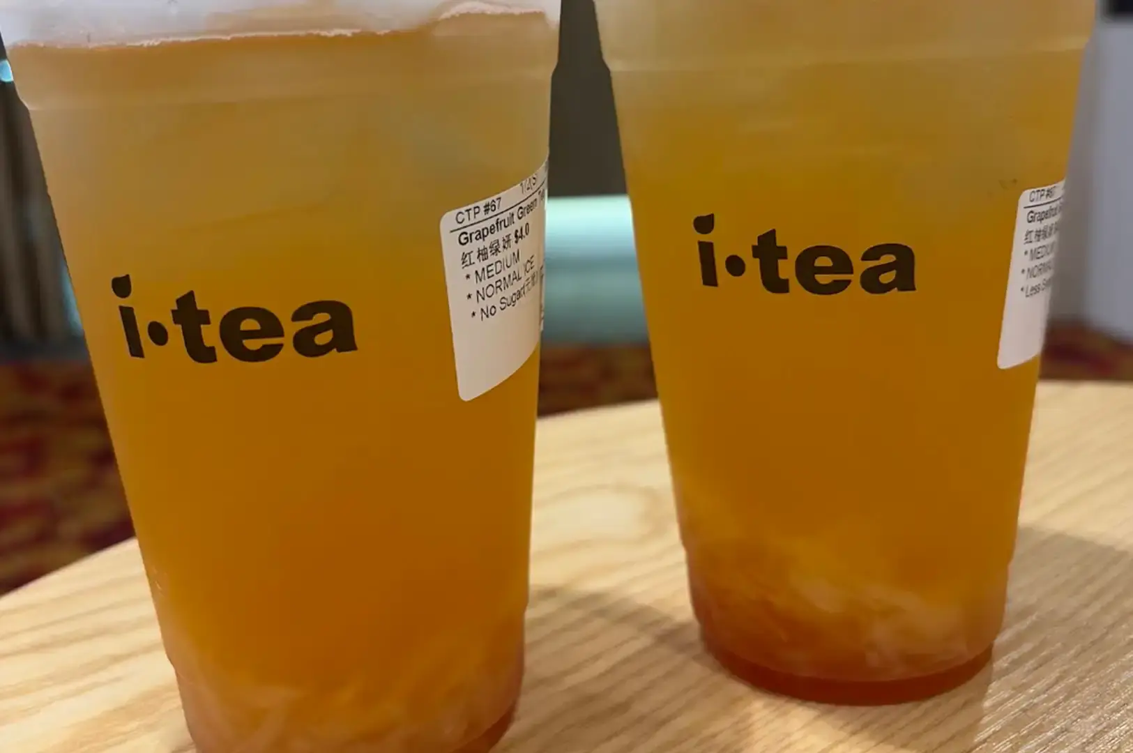 A close-up shot of two side-by-side clear plastic cups of Grapefruit Green Tea from iTea resting on a wooden tabletop. Both cups are filled with a golden-orange tea with visible grapefruit pulp settled near the bottom and bear white order stickers listing drink customizations such as sweet level and ice options.