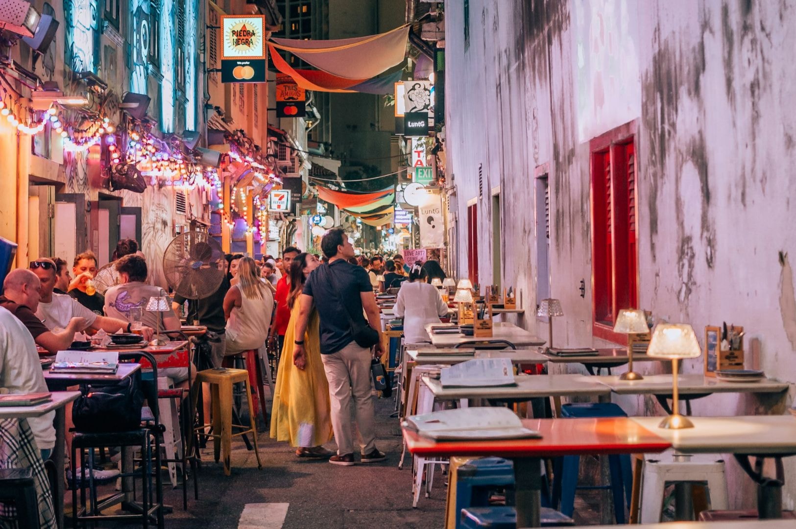 A bustling alley with colorful lights, filled with dining tables and people socializing. A vibrant, lively atmosphere with an eclectic urban feel.