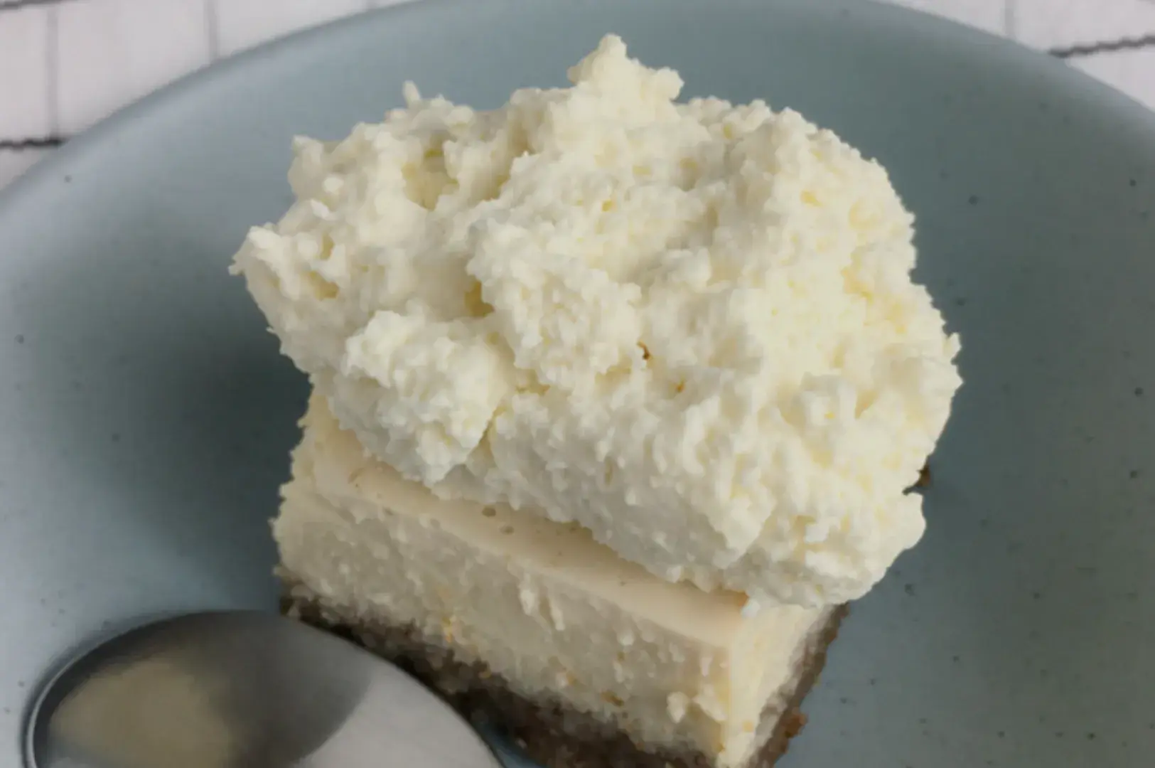 A slice of cheesecake topped with a thick layer of whipped cream on a light blue plate, with a silver spoon beside it.