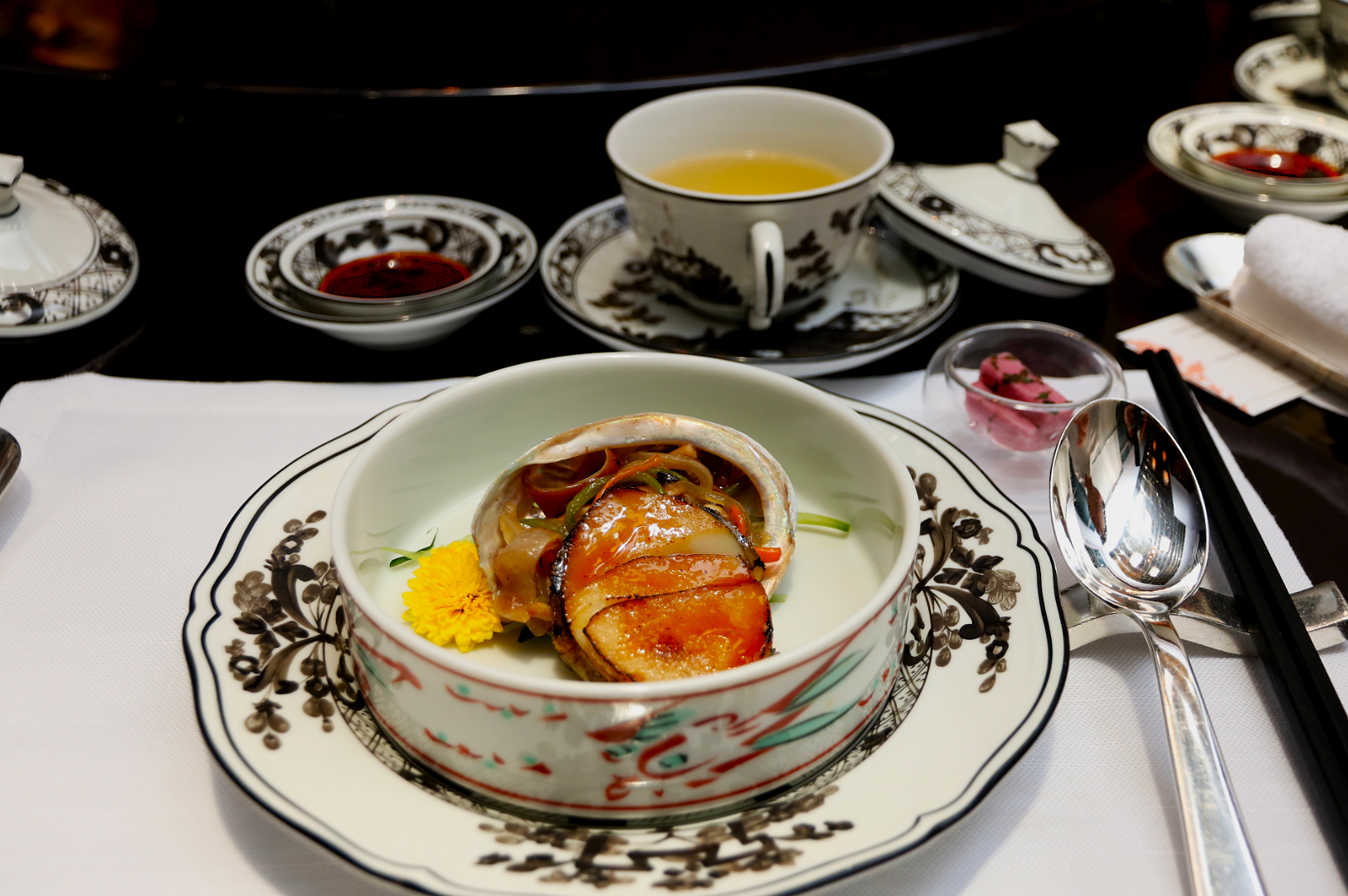 A beautifully arranged plate with abalone and vegetables on elegant floral china, accompanied by a tea cup, chopsticks, and a serene setting.