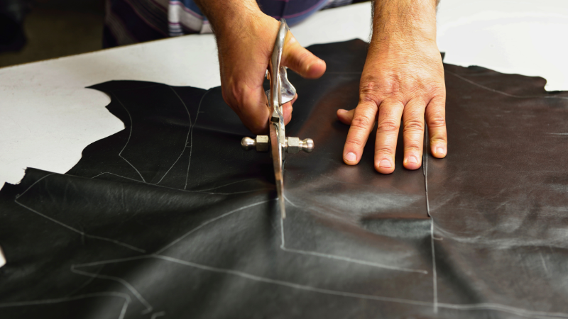 Hands use industrial scissors to cut black leather along a white chalk pattern.