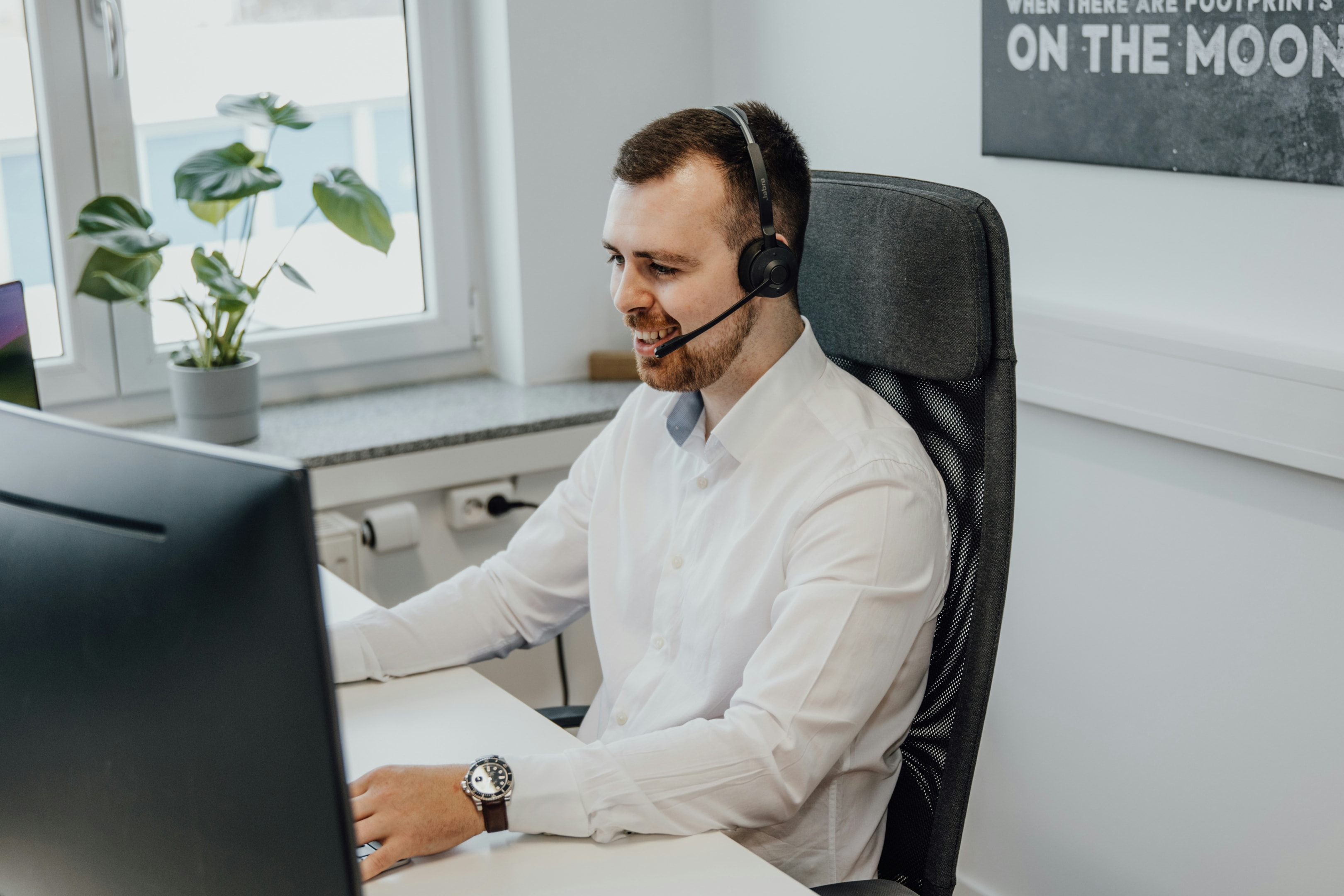 Man in a white shirt in office in front of a computer with a headset on