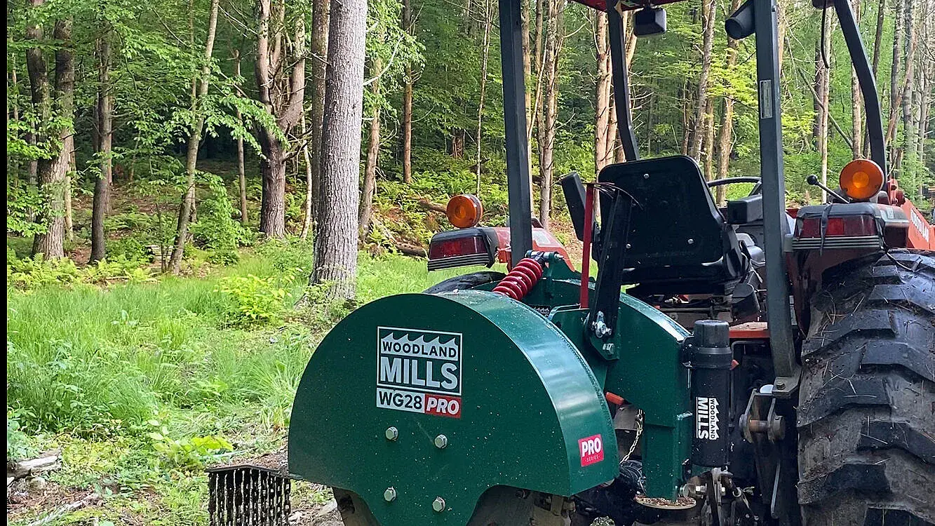 A Woodland Mills WG28 PRO Stump Grinder on the back of a tractor in a wooded area.