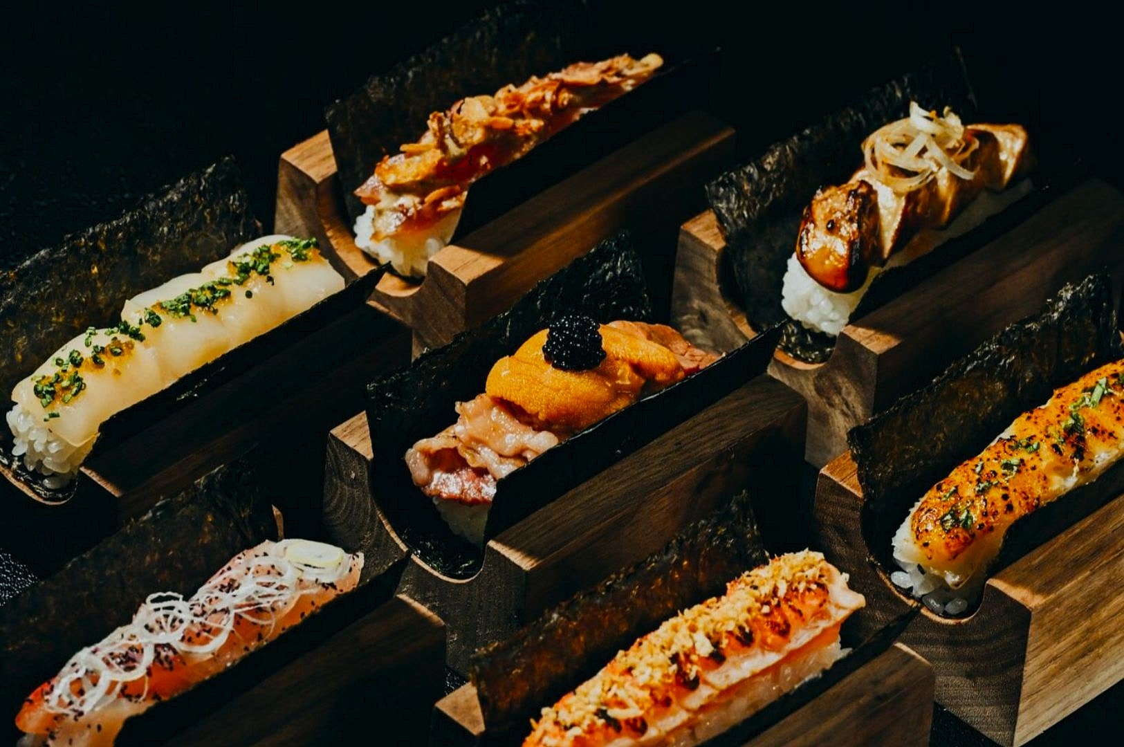 Assorted sushi pieces elegantly presented on dark wooden blocks, each topped with ingredients like seaweed, fish, and garnishes, conveying a gourmet tone.