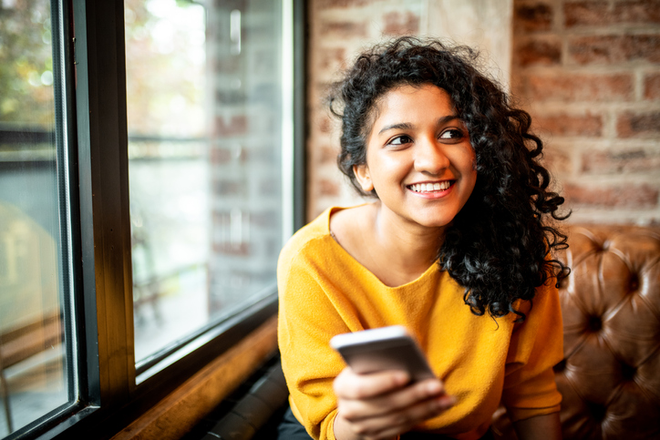 Cheerful young woman with dark curly hair in a yellow sweater holding her cell phone.