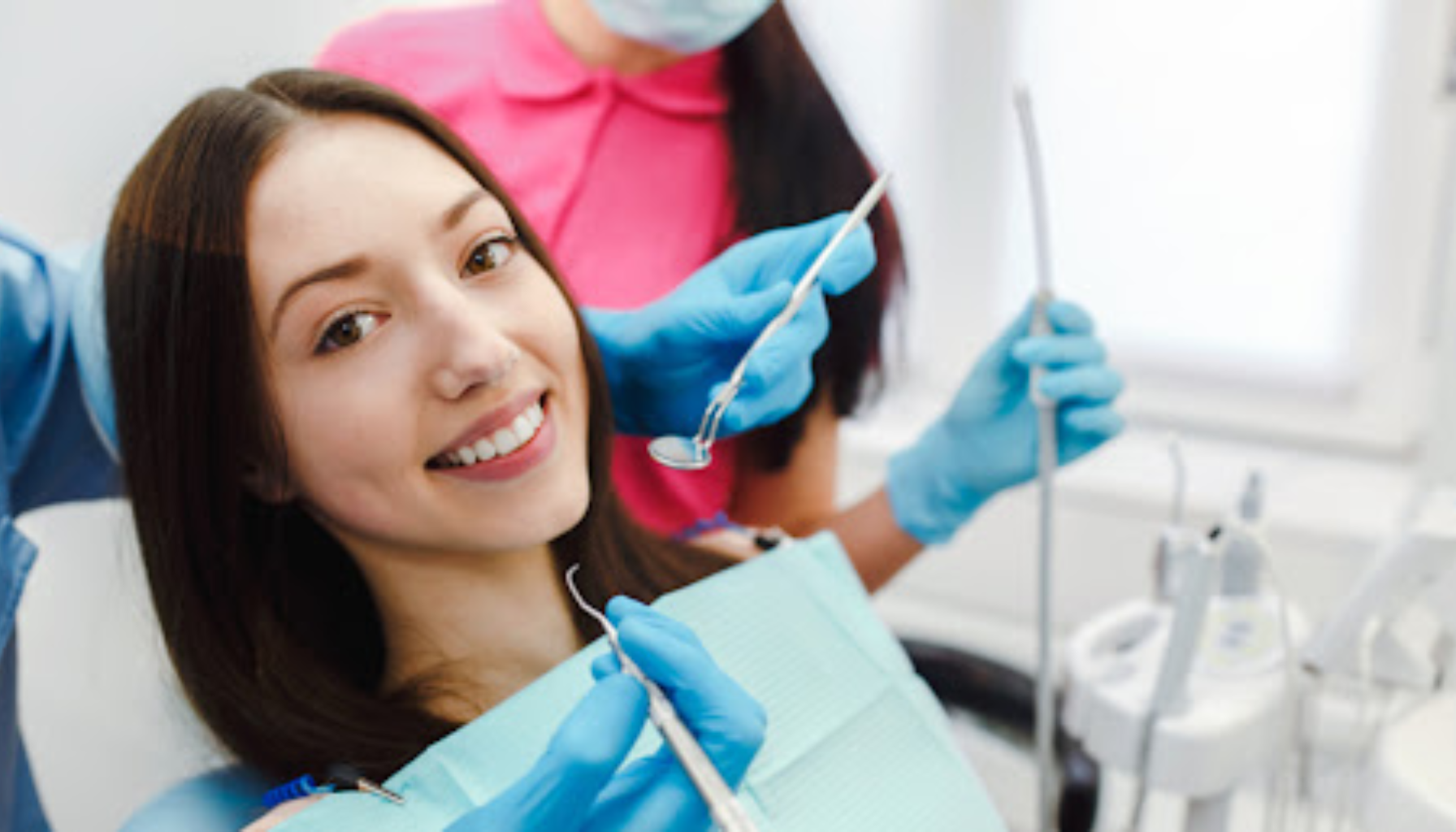 Kvinne smiler i tannlegestolen under undersøkelse, omgitt av tannhelsepersonell med hansker og munnbind i lyst behandlingsrom.