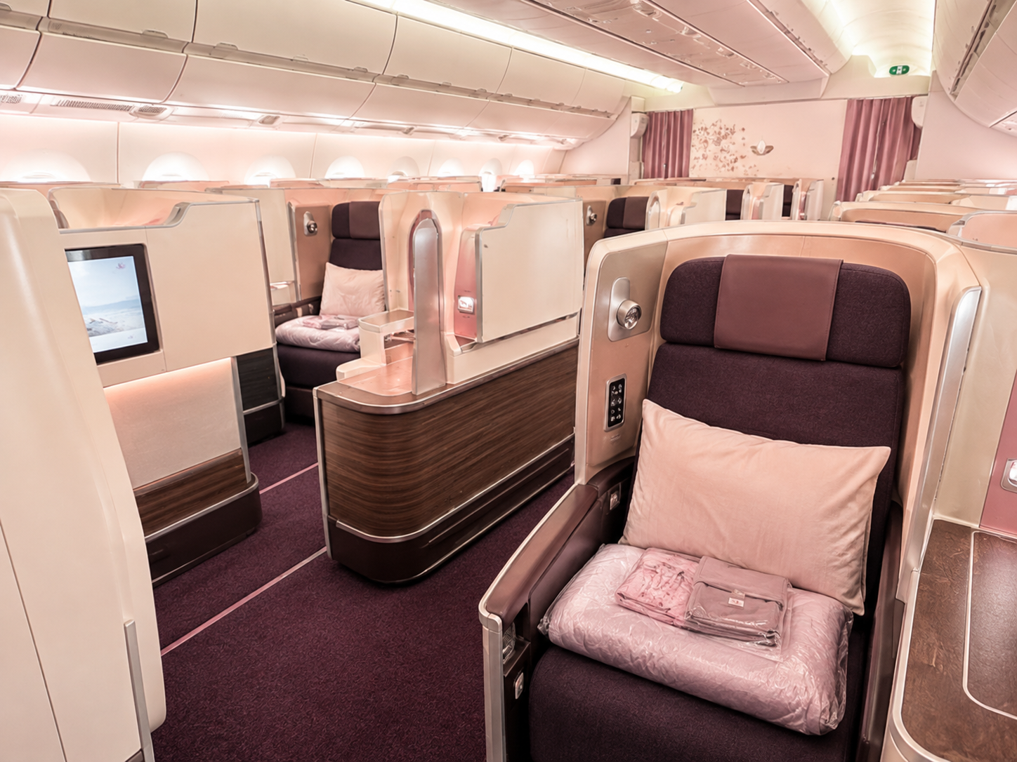 Luxury business class cabin with spacious private seats, cream partitions, dark purple upholstery, pillows and folded bedding under soft pink lighting.