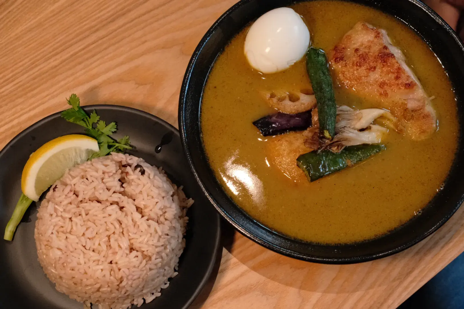 A bowl of golden curry with chicken, vegetables, and a boiled egg sits beside a mound of rice garnished with lemon and herbs on a wooden table.