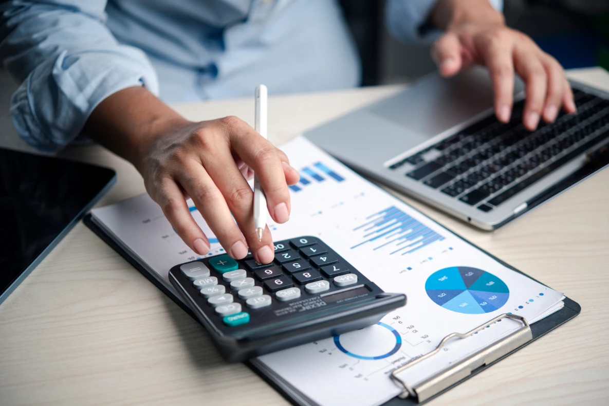 A person uses a calculator and a laptop while reviewing financial charts and graphs on a desk, illustrating what a business valuator usually does.