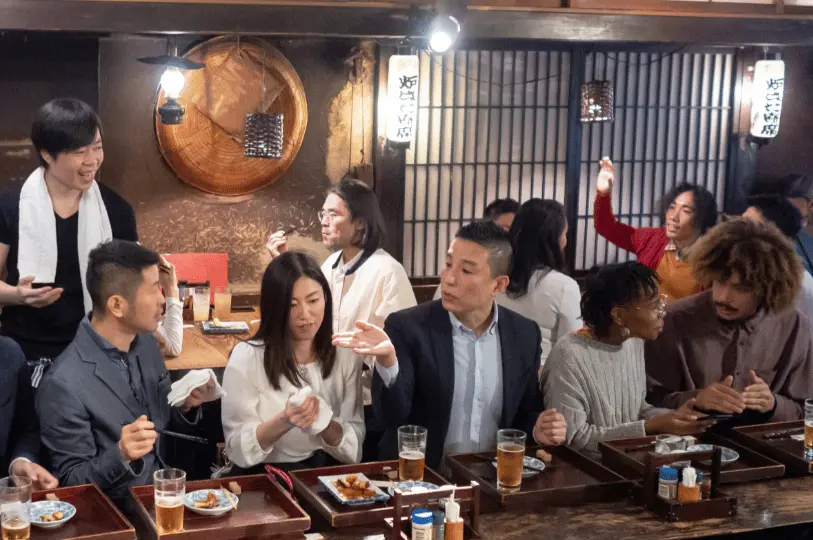 A lively scene inside a warm, wood-paneled izakaya where a chef with a towel around his neck happily interacts with a diverse group of patrons seated at the bar.
