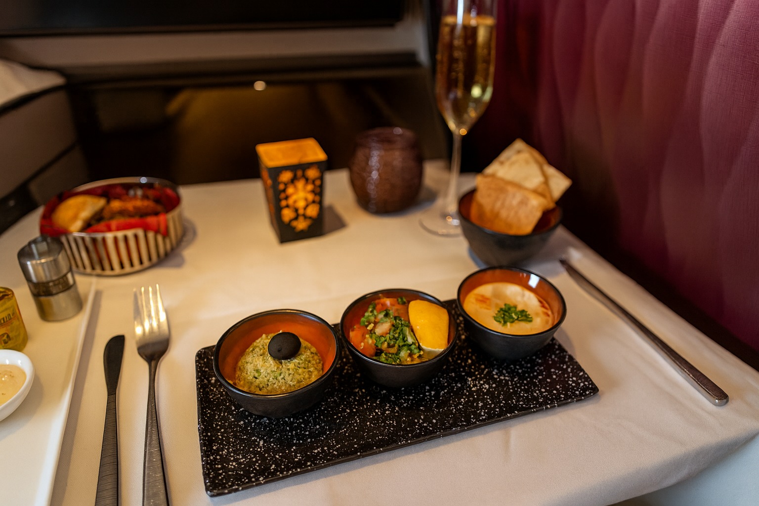 A business class in-flight dining setup with three appetizer bowls on a black speckled tray, featuring hummus, tabbouleh with lemon, and baba ghanoush topped with an olive, accompanied by pita bread, a basket of rolls, and a glass of champagne under warm cabin lighting.
