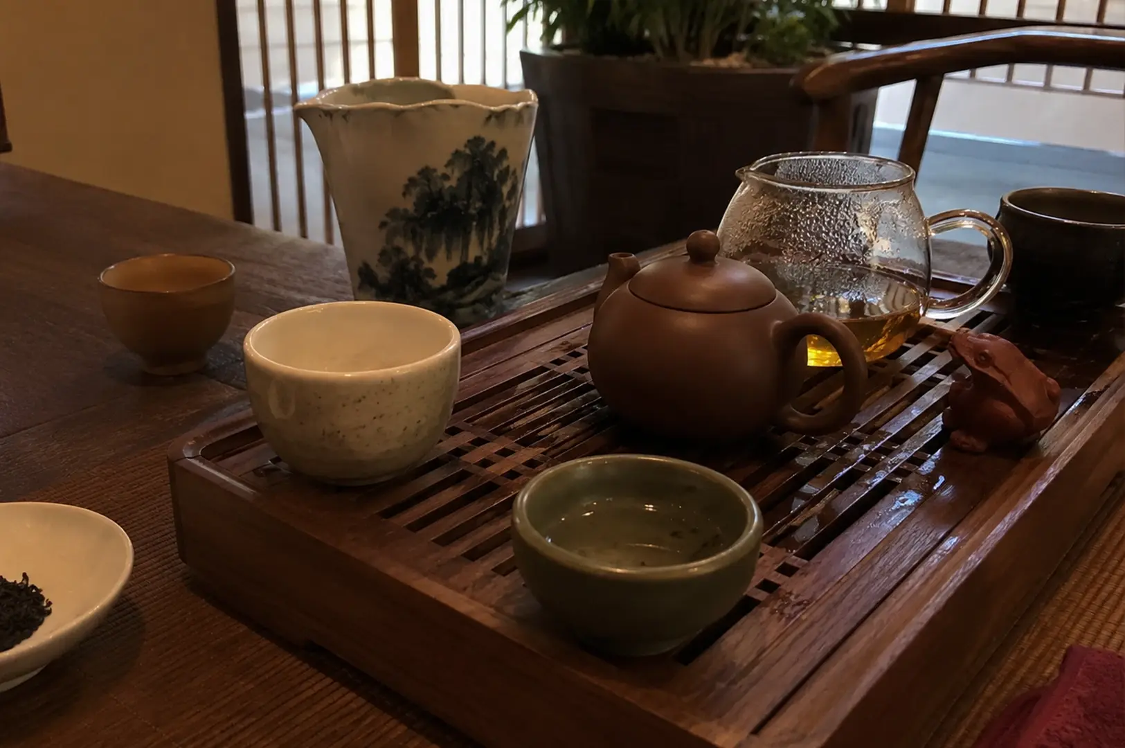 A close-up shot captures a warm, intimate tea setup resting on a dark wooden table. A rich brown clay Yixing teapot sits centrally atop a slatted wooden tea tray, accompanied by a clear glass fairness cup filled with golden tea, a small clay frog tea pet, and various ceramic teacups of different shapes and colors. In the background, a tall, white ceramic pitcher adorned with a traditional blue landscape painting stands near a window with wooden vertical blinds, while a small white dish holding dark, dry tea leaves rests on the left foreground.
