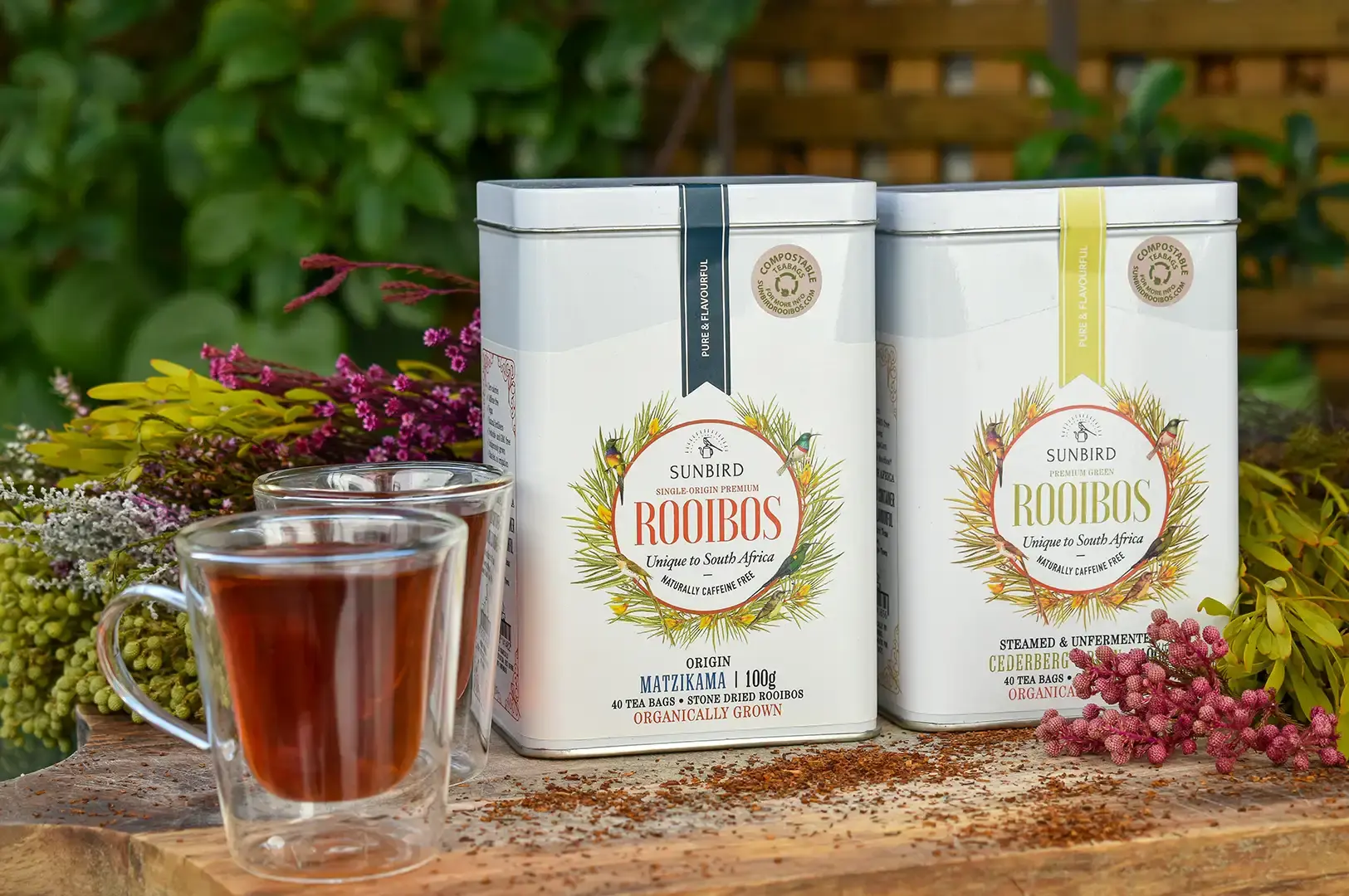 Two white Sunbird Rooibos tea tins sit on a wooden surface alongside two double-walled glass mugs filled with dark brewed tea, framed by vibrant pink, yellow, and green botanical foliage. Loose rooibos tea leaves are sprinkled across the wood, highlighting the natural, South African origin of the tea products.