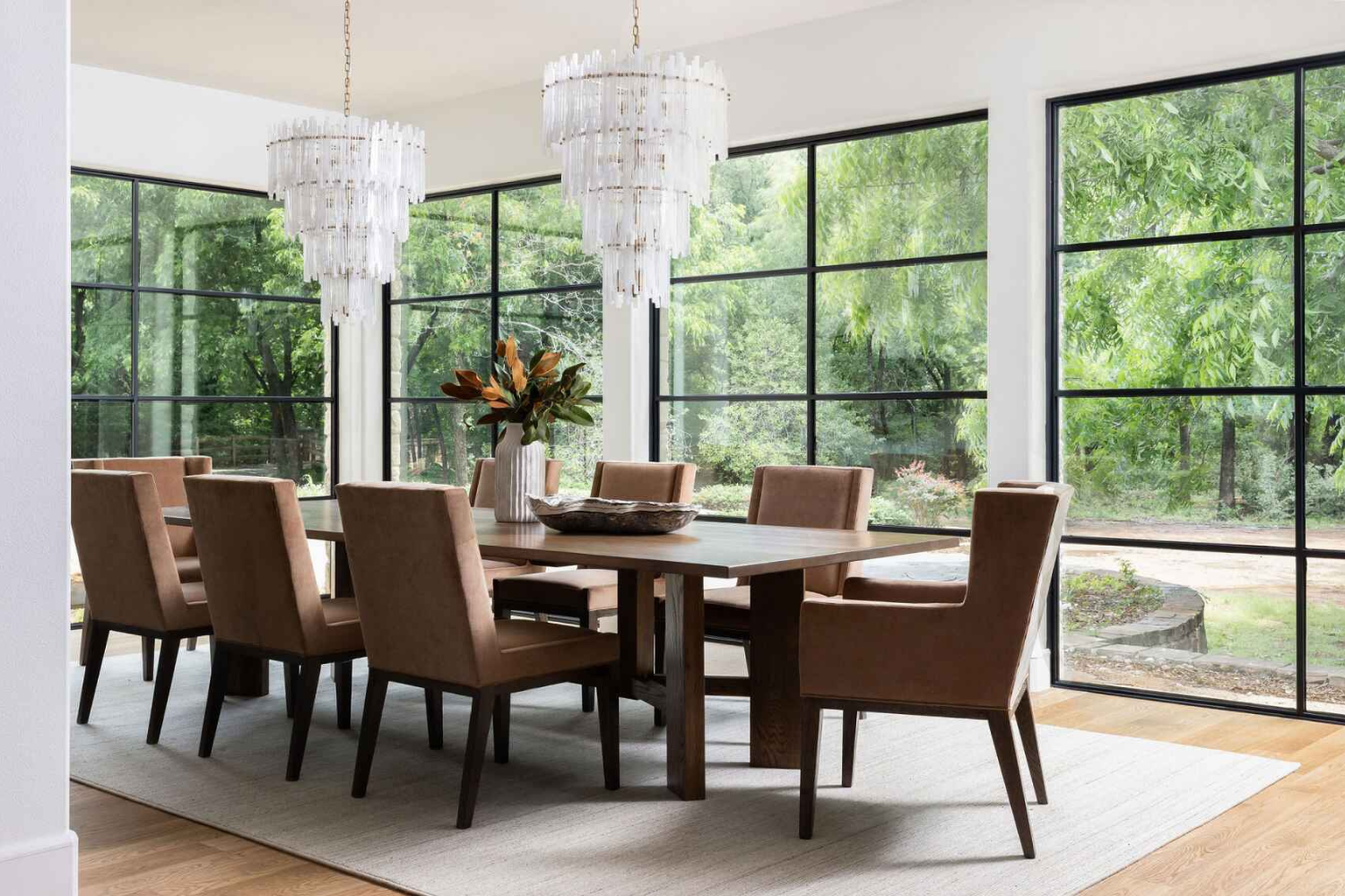 Elegant dining room with dual chandeliers, suede upholstered chairs, a wood dining table, and large black-framed windows revealing a wooded backdrop.