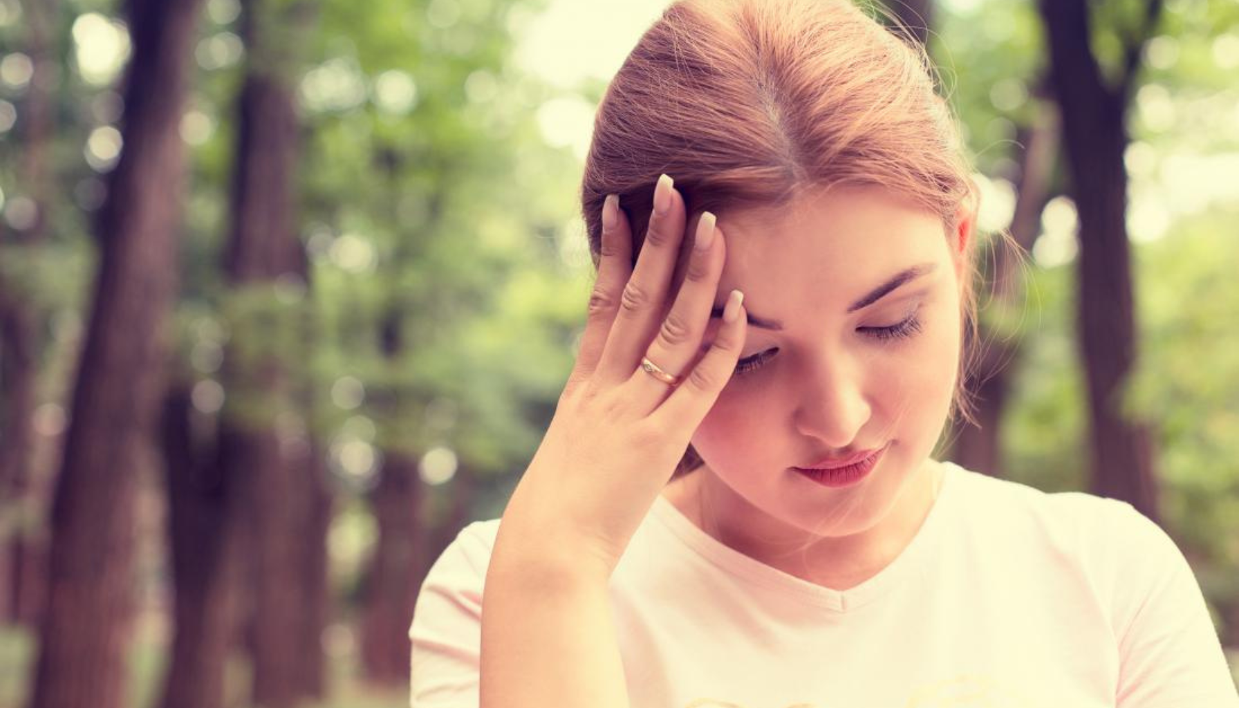 Ung kvinne utendørs holder seg til hodet, ser stresset eller svimmel ut, står blant trær i en skog.