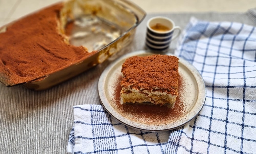 Protein tiramisu on a plate. In the background an espresso and a baking dish with tiramisu