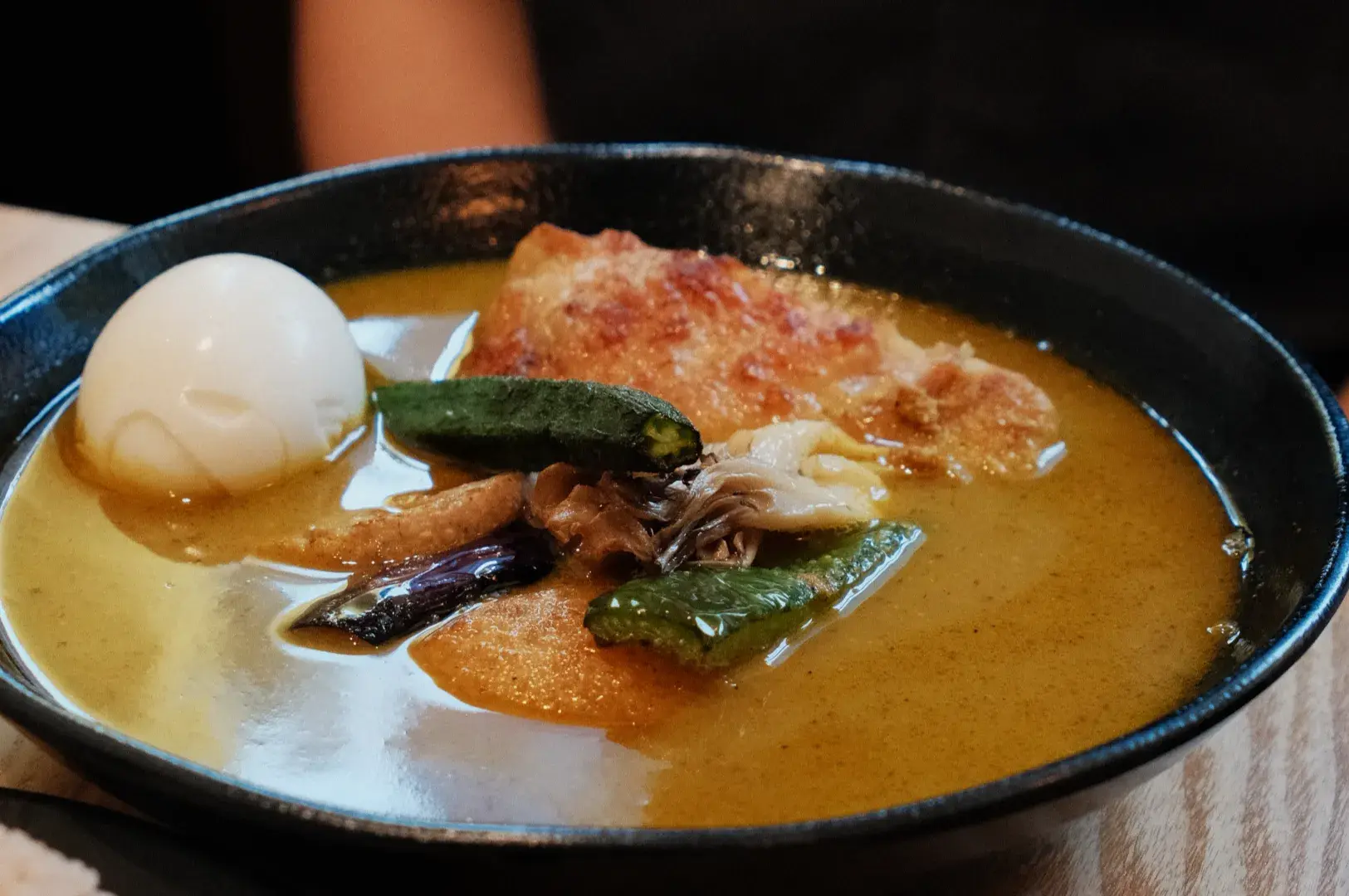 A bowl of soup curry with a soft-boiled egg, vegetables, and chicken in a rich broth. A person sits behind waiting patiently in their chair.