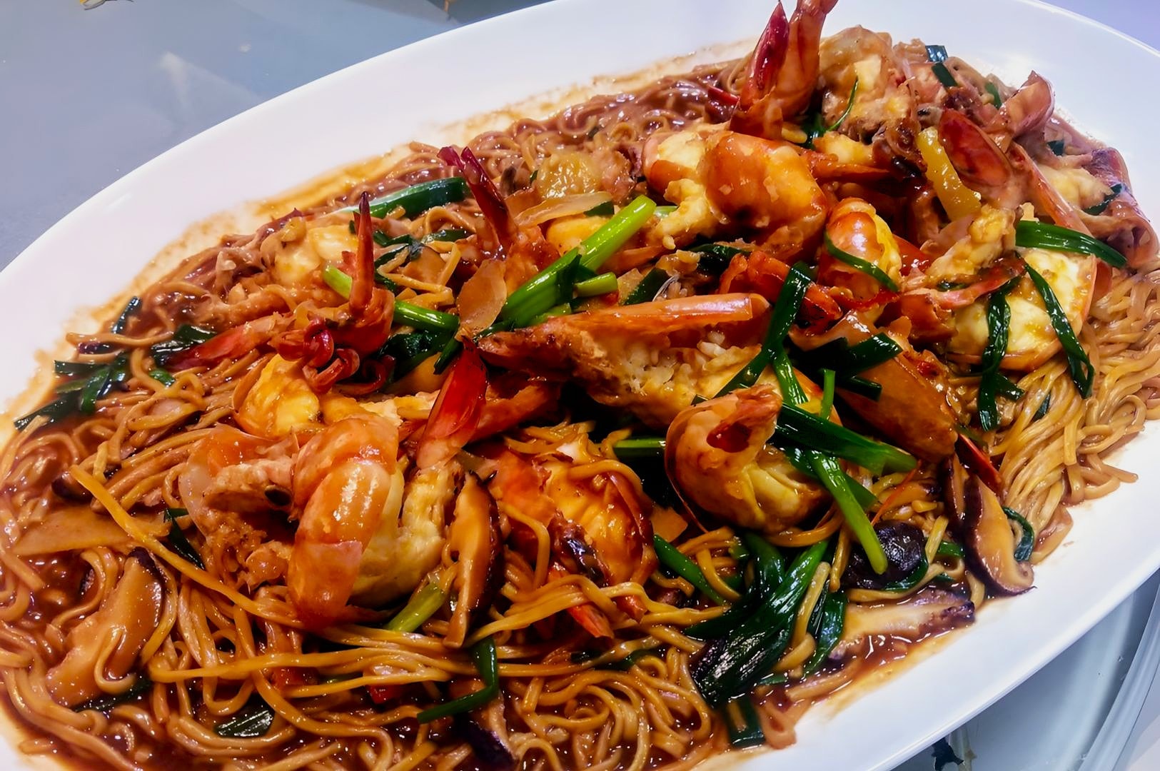 A white platter filled with stir-fried noodles topped with plump shrimp, green onions, and vegetables in a glossy brown sauce, evoking savory anticipation.