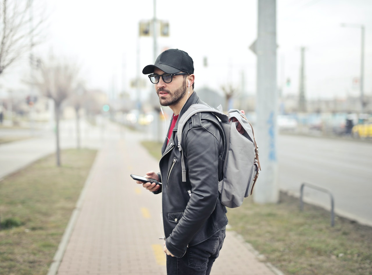 Homem de pé na rua, com o celular na mão, usando um boné, óculos e mochila nas costas - Foto: Pexels