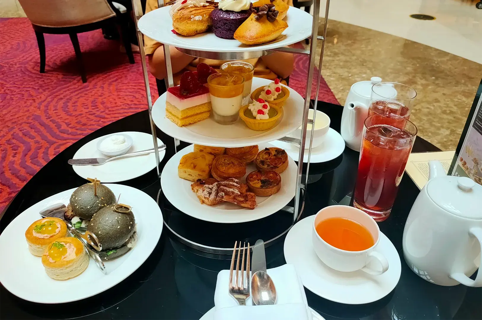 A three-tier afternoon tea stand loaded with elegant desserts, cakes, savory sliders, and mini tarts, served on a black table with hot tea.