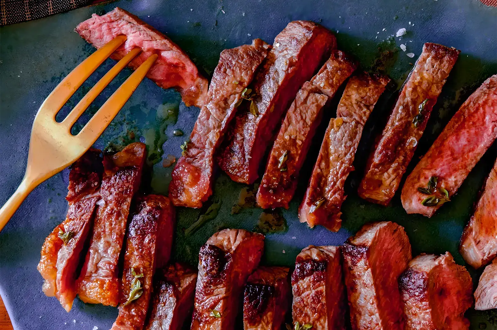 Slices of medium-rare steak are arranged on a blue plate, glistening with juices. A yellow fork holds a piece, showcasing a tender interior.