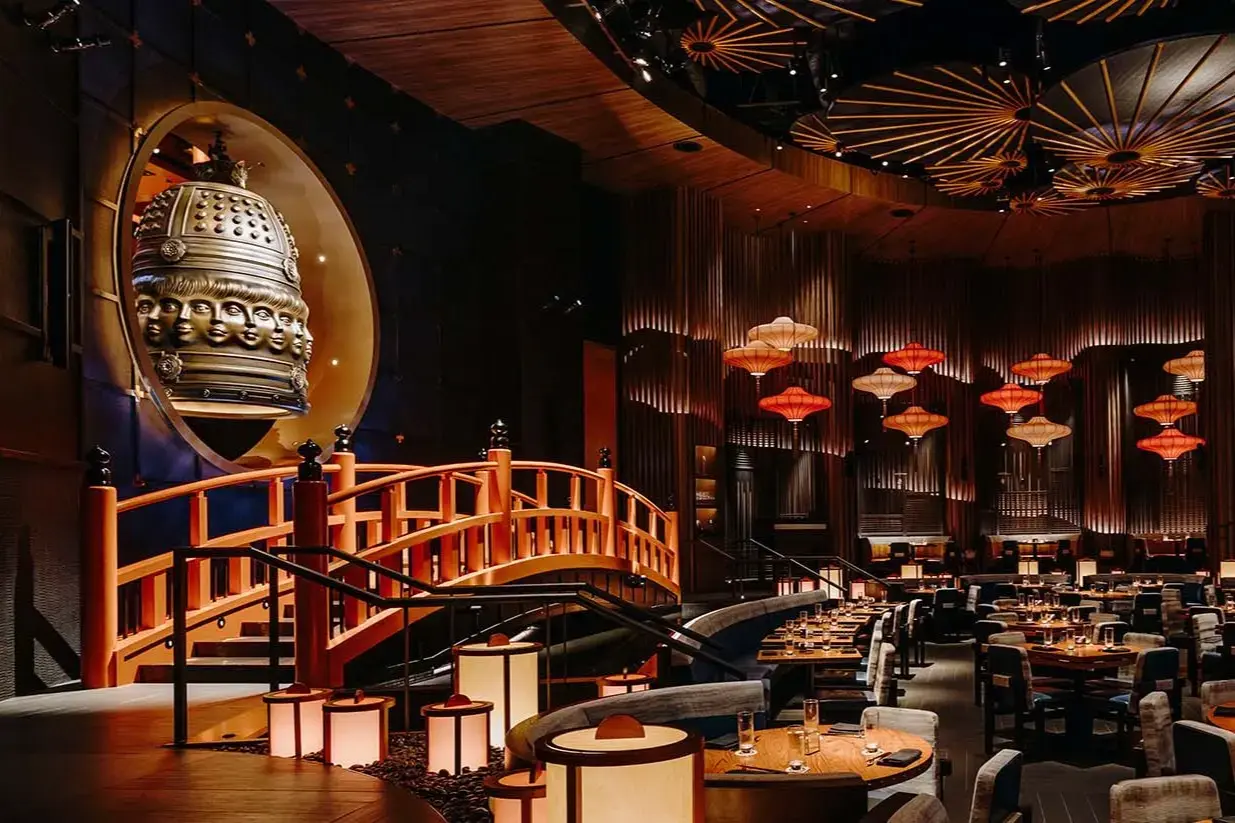 The image showcases the interior of a dimly lit, upscale restaurant featuring a striking orange wooden bridge and a large bronze bell adorned with sculpted faces. Warm glow emanates from traditional-style lanterns and hanging red lamps, illuminating the sophisticated dining area with its textured walls and circular ceiling motifs.