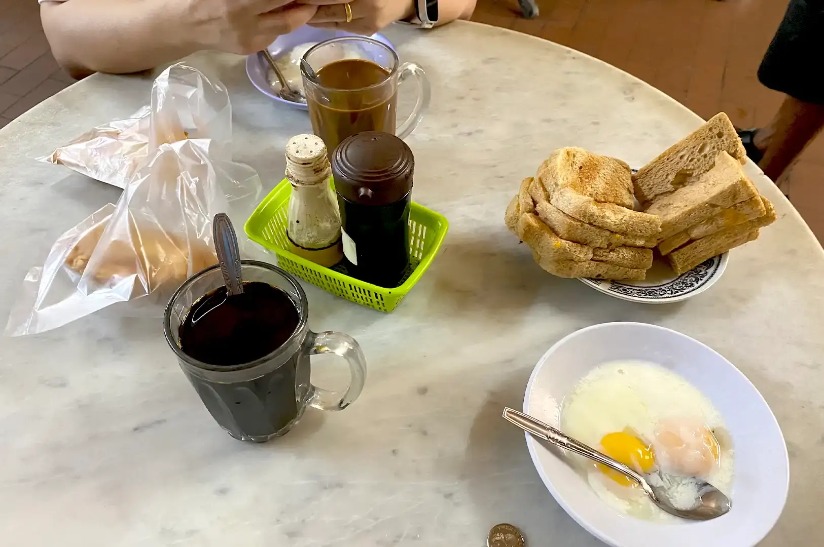 A traditional Singaporean breakfast setup featuring thick toast, soft-boiled eggs in a bowl, and classic traditional Singapore coffee in a glass mug on a marble table.