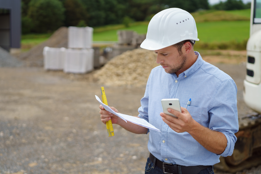 Architect or building contractor working on site standing reading a document in his hard hat with a mobile in his hand
