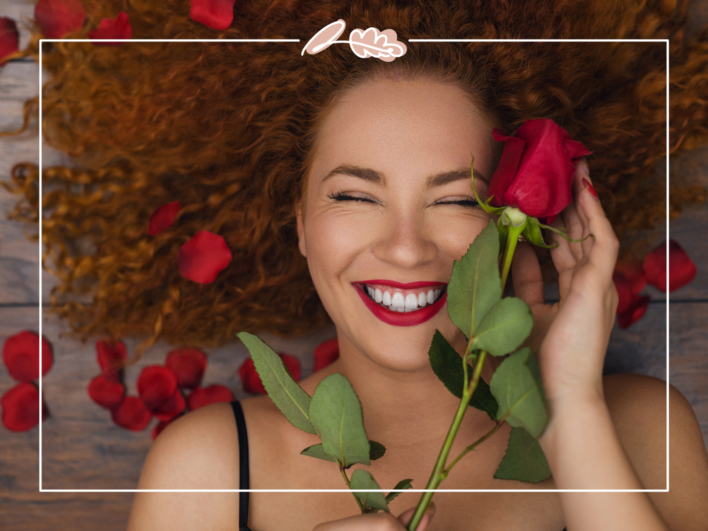 A woman with curly hair smiling joyfully, holding a red rose near her face, surrounded by scattered rose petals on the ground. Fabulous Flowers & Gifts